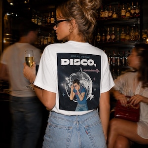 May include: A white short-sleeved t-shirt featuring a rectangular graphic on the back. The graphic displays a disco ball, stars, and a person wearing sunglasses, with the text "KISS ALL THE TIME DISCO, occasionally." printed in white and red.