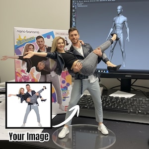 May include: A figurine of a man holding a woman, both in casual attire, with a box in the background. The man wears a black leather jacket, gray pants, and white sneakers. The woman wears a black top, gray jeans, and black heels. A computer monitor displays a 3D model.