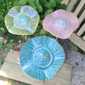 Peut inclure: Trois tuteurs de jardin en céramique en forme de fleurs avec des finitions brillantes. Une fleur est vert clair avec un centre bleu, une est rose pâle avec un centre blanc texturé, et une est bleu vif avec un centre tacheté. Bords ondulés.