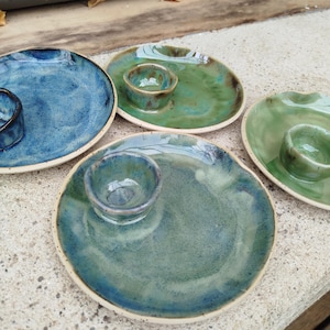 May include: A collection of handmade ceramic serving dishes in shades of blue and green. Each dish features a small, attached bowl, perfect for dips or sauces. The plates have a rustic, textured appearance with a glossy finish.