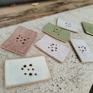 Könnte beinhalten: Eine Sammlung rechteckiger Keramik-Seifenschalen in verschiedenen Farben, darunter Rosa, Grün und Weiß. Jede Schale hat eine Reihe kreisförmiger Abflusslöcher. Die Schalen sind auf einer Betonoberfläche angeordnet.