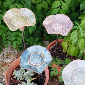 Peut inclure: Quatre tuteurs de jardin en céramique en forme de fleur avec des bords ondulés et des centres texturés. Les pétales présentent des émaux irisés brillants dans des tons vert pâle, rose doux, bleu clair et blanc cassé, montés sur une tige métallique.