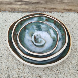 Może przedstawiać: Zestaw trzech ceramicznych misek o różnych rozmiarach, umieszczonych jedna w drugiej. Miski mają w środku turkusowo-biały wzór w wiry, z brązową obwódką i białym zewnętrznym brzegiem. Miski są ułożone na betonowej powierzchni.