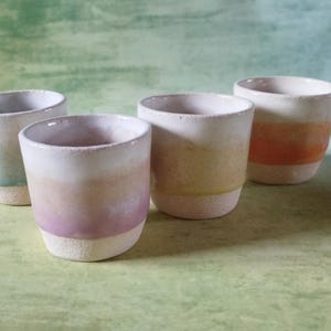 Könnte beinhalten: Vier handgefertigte Keramikbecher mit Farbverlauf. Jeder Becher hat eine einzigartige Mischung aus Farben, darunter Türkis, Rosa, Gelb und Orange, mit einem weißen Rand und einem strukturierten Boden. Die Becher sind auf einer grünen Oberfläche angeordnet.