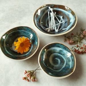 May include: Three handmade ceramic bowls with a teal and brown glaze. One bowl contains white cotton swabs, another has an orange flower, and the third is empty. The bowls are arranged on a light surface.
