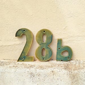Op de afbeelding: Keramische huisnummers, met '28b'. De cijfers zijn teal en olijfgroen, met montagegaten. De achtergrond is een getextureerde, lichtbeige muur. Perfect voor een huis.