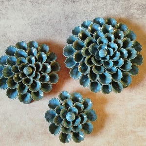 Pode incluir: Três esculturas de parede de cerâmica circulares em forma de flores desabrochando com pétalas em camadas. Cada peça apresenta um esmalte brilhante e variado em tons de azul-petróleo, azul e verde, com um centro mais escuro.