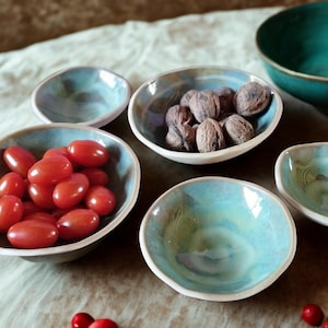 Op de afbeelding: Een verzameling handgemaakte keramische kommen in verschillende maten en vormen. De kommen hebben een turquoise en witte glazuur, sommige gevuld met rode tomaten en walnoten. De kommen zijn gerangschikt op een neutraal gekleurd oppervlak.