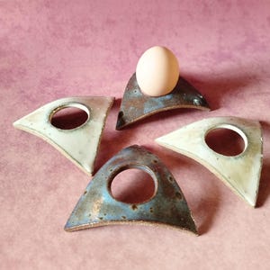 Pode incluir: Quatro suportes para ovos em cerâmica de formato triangular dispostos em círculo. Dois suportes apresentam um esmalte brilhante em tom branco sujo, enquanto os outros dois exibem um acabamento metálico manchado em azul e castanho.