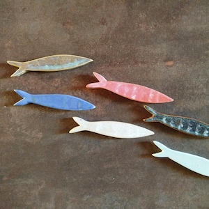 May include: A collection of six ceramic fish-shaped ornaments in various colors including tan, blue, pink, white, and a mottled dark grey. Each piece features a smooth, elongated body with a distinct tail fin, arranged in a scattered pattern.