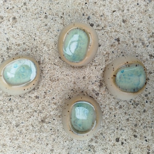 Peut inclure: Quatre pièces en céramique faites à la main, beige clair à l'extérieur et turquoise à l'intérieur. Chaque pièce est arrondie avec un bord surélevé et de petits points noirs. Elles sont disposées sur une surface texturée gris clair.