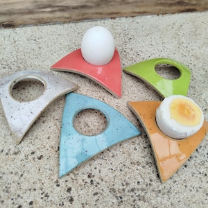 Puede incluir: Colección de coloridos hueveros de cerámica en rojo, verde, azul, naranja y blanco moteado. Cada soporte triangular tiene una abertura circular y sostiene un huevo entero o a la mitad. Los soportes están sobre una superficie de hormigón.