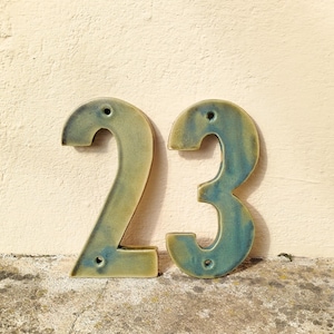 May include: Two ceramic house numbers, 2 and 3, in a teal and green glaze. The numbers have a glossy finish and are mounted on a light beige wall. Each number has pre-drilled holes for mounting.