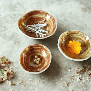Puede incluir: Tres cuencos de cerámica pequeños, de forma orgánica, con un patrón de remolino marrón y crema. Uno contiene bastoncillos de algodón, otro perlas y el tercero una flor de color naranja brillante. Los cuencos están dispuestos sobre una superficie neutra.