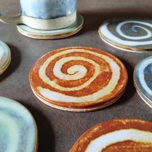 May include: A selection of handmade ceramic coasters with a spiral pattern. The coasters display shades of orange, white, blue, and grey. A blue and white mug rests on a matching coaster. These coasters are ideal for protecting surfaces from hot or cold beverages.