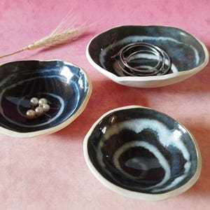 Può includere: Tre ciotole in ceramica fatte a mano con un design a vortice blu scuro e bianco. Le ciotole hanno un bordo bianco e contengono gioielli e perle. Le ciotole sono su una superficie rosa.
