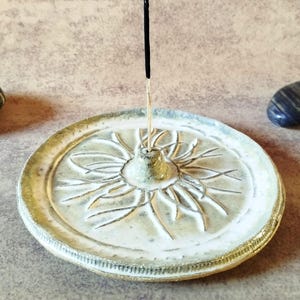 May include: A round ceramic incense holder with a white and pale green glaze featuring a carved radial floral pattern. A small central cone holds a single black incense stick upright. The edges of the dish have a textured, metallic gold finish.