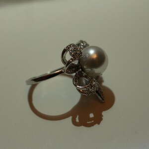 Puede incluir: Un anillo plateado con una perla grande y redonda como punto focal. El dise&ntilde;o del anillo incluye detalles intrincados y brillantes alrededor de la perla, realzando su atractivo. El anillo se presenta sobre un fondo liso.