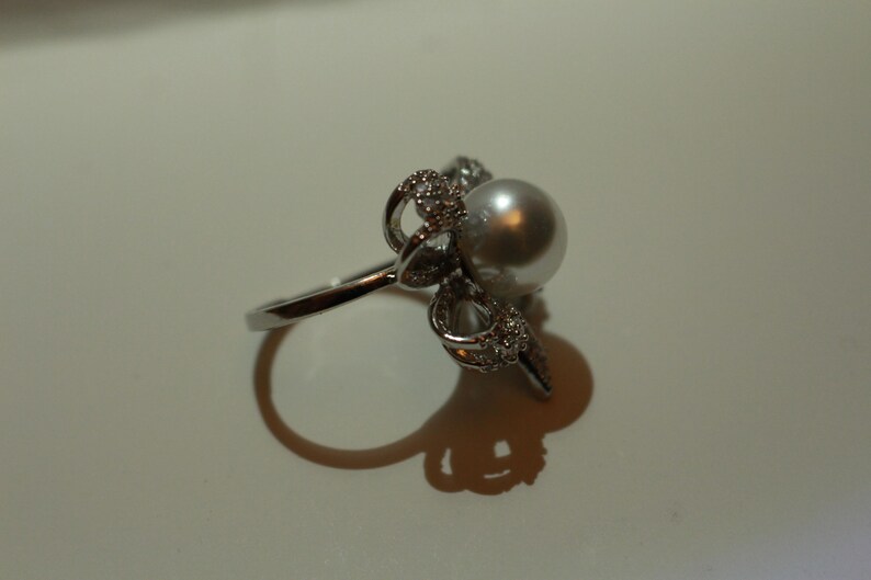 Puede incluir: Un anillo plateado con una perla grande y redonda como punto focal. El dise&ntilde;o del anillo incluye detalles intrincados y brillantes alrededor de la perla, realzando su atractivo. El anillo se presenta sobre un fondo liso.