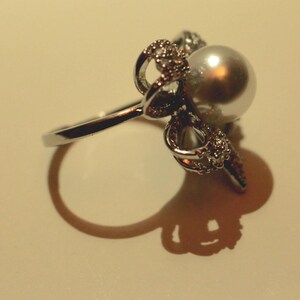 Puede incluir: Un anillo plateado con una gran perla brillante como pieza central. El anillo est&aacute; adornado con intrincados y brillantes adornos alrededor de la perla, a&ntilde;adiendo un toque de elegancia. El dise&ntilde;o del anillo se destaca por su sombra.