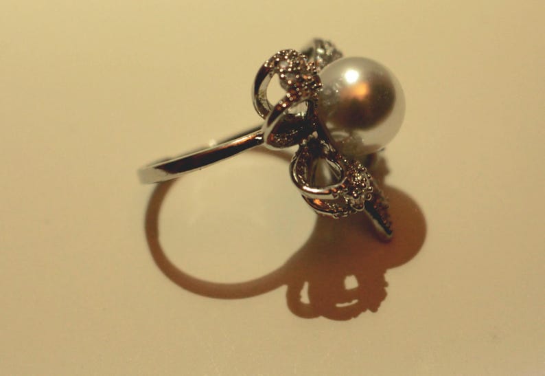 Puede incluir: Un anillo plateado con una gran perla brillante como pieza central. El anillo est&aacute; adornado con intrincados y brillantes adornos alrededor de la perla, a&ntilde;adiendo un toque de elegancia. El dise&ntilde;o del anillo se destaca por su sombra.