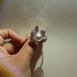 Puede incluir: Anillo plateado con una perla grande y redonda en el centro, rodeada por un dise&ntilde;o floral. Los p&eacute;talos est&aacute;n adornados con peque&ntilde;os cristales brillantes y detalles m&aacute;s oscuros. El anillo se muestra sobre un fondo neutro.