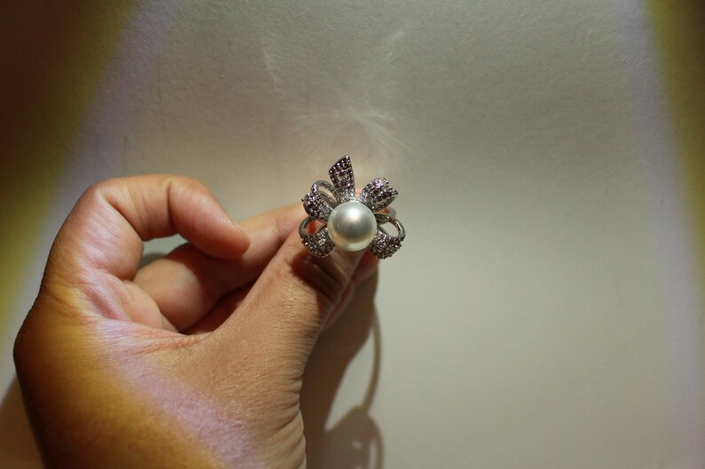 Puede incluir: Anillo plateado con una perla grande y redonda en el centro, rodeada por un dise&ntilde;o floral. Los p&eacute;talos est&aacute;n adornados con peque&ntilde;os cristales brillantes y detalles m&aacute;s oscuros. El anillo se muestra sobre un fondo neutro.