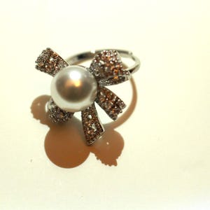Puede incluir: Un anillo plateado con una gran piedra central redonda con forma de perla. El anillo presenta un dise&ntilde;o en forma de lazo, adornado con peque&ntilde;os cristales brillantes. El anillo est&aacute; sobre un fondo claro.
