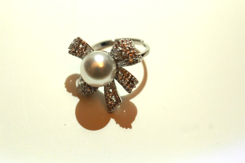 Puede incluir: Un anillo plateado con una gran piedra central redonda con forma de perla. El anillo presenta un dise&ntilde;o en forma de lazo, adornado con peque&ntilde;os cristales brillantes. El anillo est&aacute; sobre un fondo claro.