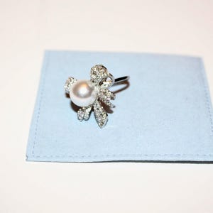 Puede incluir: Un anillo plateado con una perla blanca redonda y grande en el centro, rodeada por un dise&ntilde;o floral adornado con peque&ntilde;os cristales brillantes. El anillo descansa sobre una tela cuadrada azul claro.