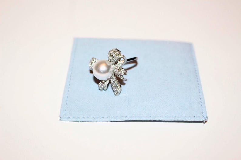 Puede incluir: Un anillo plateado con una perla blanca redonda y grande en el centro, rodeada por un dise&ntilde;o floral adornado con peque&ntilde;os cristales brillantes. El anillo descansa sobre una tela cuadrada azul claro.