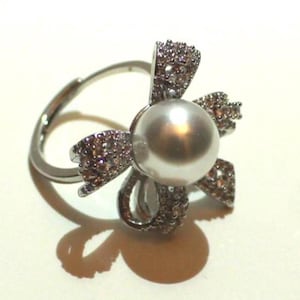 Puede incluir: Un anillo plateado con una perla gris redonda y grande en el centro, rodeada por un dise&ntilde;o en forma de flor incrustado con peque&ntilde;os cristales brillantes. El anillo tiene una banda ajustable.