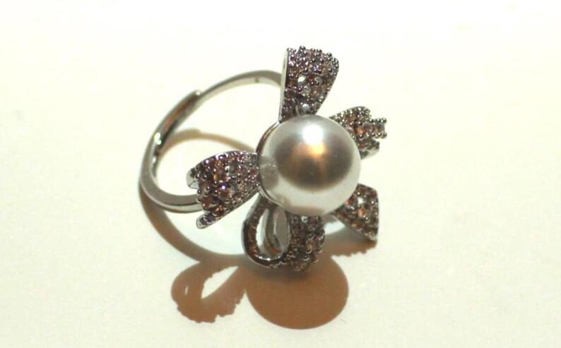 Puede incluir: Un anillo plateado con una perla gris redonda y grande en el centro, rodeada por un dise&ntilde;o en forma de flor incrustado con peque&ntilde;os cristales brillantes. El anillo tiene una banda ajustable.