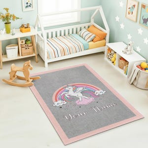 Può includere: Un tappeto rettangolare grigio con bordo rosa presenta un disegno di unicorno. L'unicorno è bianco con un arcobaleno, nuvole e la scritta "Your Name". Il tappeto si trova nella cameretta di un bambino con un letto bianco, un tavolo e un cavallo a dondolo.