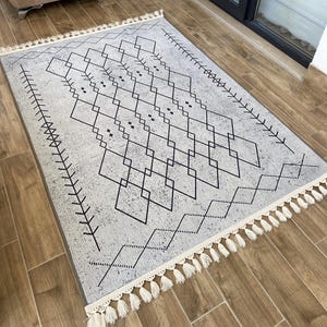 Könnte beinhalten: Ein rechteckiger grauer Teppich mit einem geometrischen schwarzen Design und weißen Fransen. Der Teppich weist ein sich wiederholendes Muster aus Rauten, Linien und Pfeilen auf. Der Teppich liegt auf einem Holzboden.