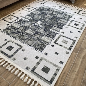 Peut inclure: Un tapis rectangulaire avec un fond crème et un motif géométrique en nuances de gris et de noir. Le tapis présente un motif central avec des éléments floraux et abstraits, entouré de formes carrées et rectangulaires. Des glands blancs ornent les bords.