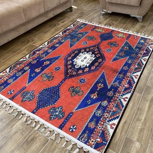 Può includere: Un tappeto rettangolare con un vivace sfondo rosso e intricati motivi geometrici blu e bianchi. Il tappeto presenta un motivo a medaglione centrale e un bordo con frange bianche. Misura circa 152 cm x 213 cm.