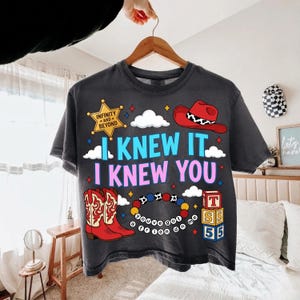 K&ouml;nnte beinhalten: Ein anthrazitfarbenes T-Shirt mit einem Grafikdruck, der einen gelben Sheriffstern mit der Aufschrift Infinity and Beyond, einen roten Cowboyhut, rote Cowboystiefel, ein buntes Perlenarmband und einen Stapel Alphabetw&uuml;rfel mit T, S, 5, 5 zeigt.