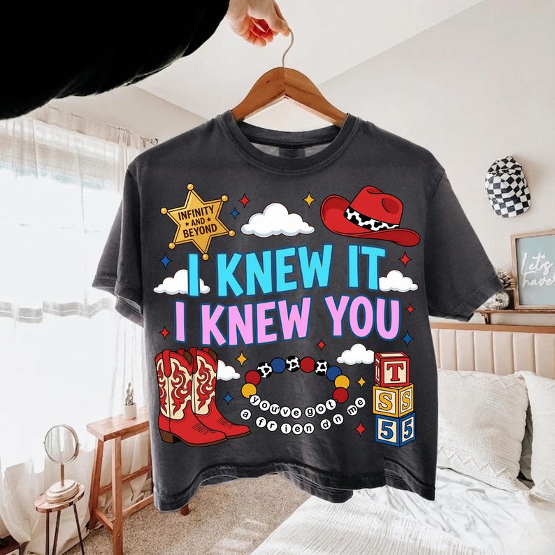 K&ouml;nnte beinhalten: Ein anthrazitfarbenes T-Shirt mit einem Grafikdruck, der einen gelben Sheriffstern mit der Aufschrift Infinity and Beyond, einen roten Cowboyhut, rote Cowboystiefel, ein buntes Perlenarmband und einen Stapel Alphabetw&uuml;rfel mit T, S, 5, 5 zeigt.