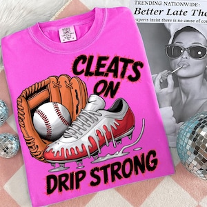 May include: A pink t-shirt with the words "CLEATS ON DRIP STRONG" in black. The shirt features a baseball glove, a baseball, and a red and white baseball cleat with dripping accents. The shirt also has a small tag.