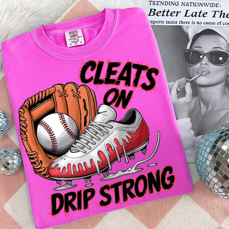 May include: A pink t-shirt with the words "CLEATS ON DRIP STRONG" in black. The shirt features a baseball glove, a baseball, and a red and white baseball cleat with dripping accents. The shirt also has a small tag.