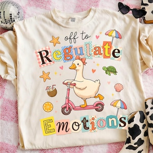 Puede incluir: Camiseta color crema con un gráfico caprichoso de un pato montando un scooter rosa, rodeado de estrellas de mar, corazones y una tortuga. El texto "off to Regulate Emotions" se muestra en letras coloridas y juguetonas. Un diseño divertido y alegre.