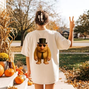 May include: Cream-colored t-shirt featuring a cartoon troll wearing a top hat, sunglasses, and a black tie. The image is on the back of the shirt. The person is standing outside on a fall day with pumpkins and corn stalks.