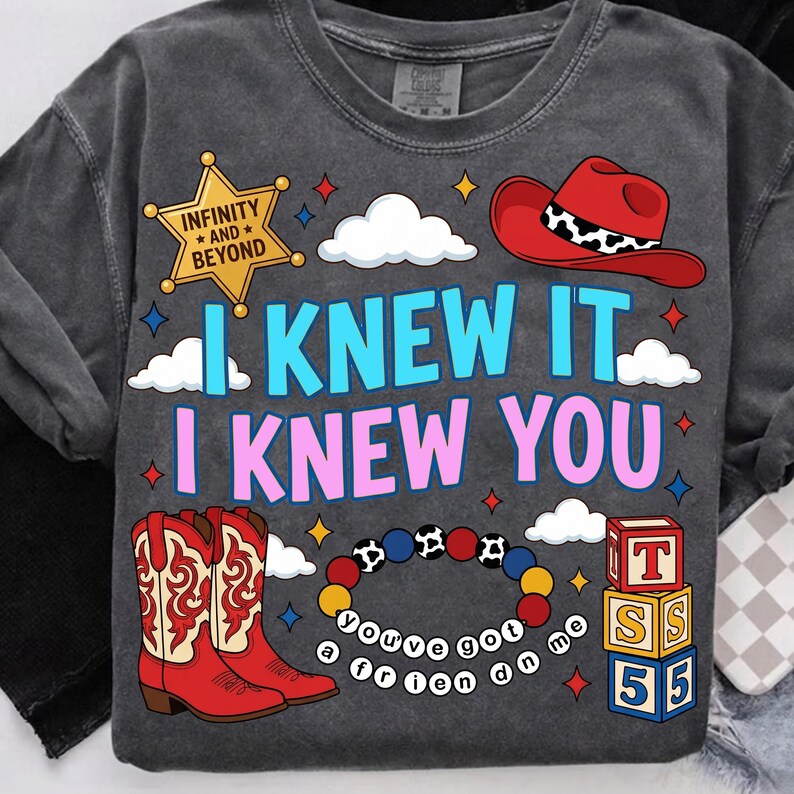 K&ouml;nnte beinhalten: Ein anthrazitfarbenes T-Shirt mit einem Grafikdruck und dem Text "I KNEW IT I KNEW YOU" in Blau und Pink. Das Design zeigt einen gelben Sheriffstern mit "INFINITY AND BEYOND", einen roten Cowboyhut, rote Stiefel, ein Perlenarmband und Spielzeugw&uuml;rfel.