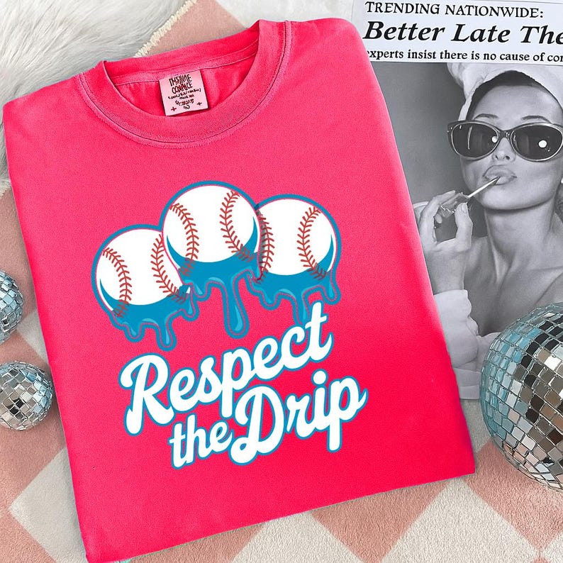 May include: A bright pink t-shirt featuring a baseball-themed graphic. The design includes three baseballs with blue dripping accents and the text "Respect the Drip" in a stylized font. The shirt has a crew neck and short sleeves.