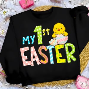 May include: Black sweatshirt with the text "My 1st Easter" in colorful letters. A yellow chick is hatching from an egg. The letters have floral and polka dot patterns, with a bunny design on the letter "E".
