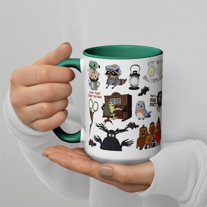 May include: White ceramic mug with a green handle and interior. The mug features cartoon illustrations of a raccoon, frog, and lantern. Text includes "Ain't that just the way" and "You make beautiful things". A fun and quirky design.