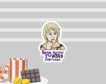 Peut inclure: Un autocollant avec une illustration de dessin animé d'une femme aux cheveux blonds, portant un haut beige et violet, et une main sur le menton. Le texte "Being Normal is VASTLY Overrated" est en violet et bleu.