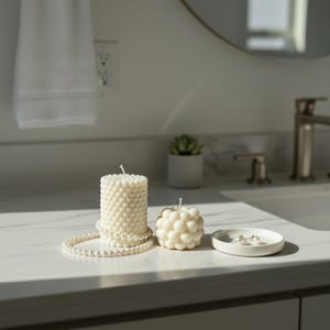 Puede incluir: Dos velas de color marfil con texturas únicas se encuentran sobre una encimera blanca. Una vela es cilíndrica con una superficie con forma de burbujas, la otra es esférica. También hay un collar de perlas y un plato blanco pequeño con pendientes.