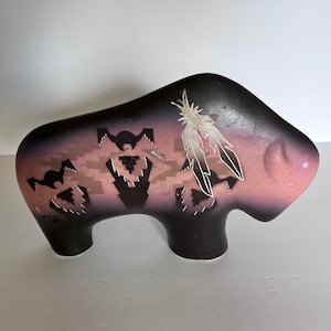 Puede incluir: Una escultura de bisonte de cerámica en tonos negros, morados y rosas. El bisonte presenta diseños geométricos recortados y detalles de plumas blancas. La escultura tiene un acabado liso y es un objeto decorativo.